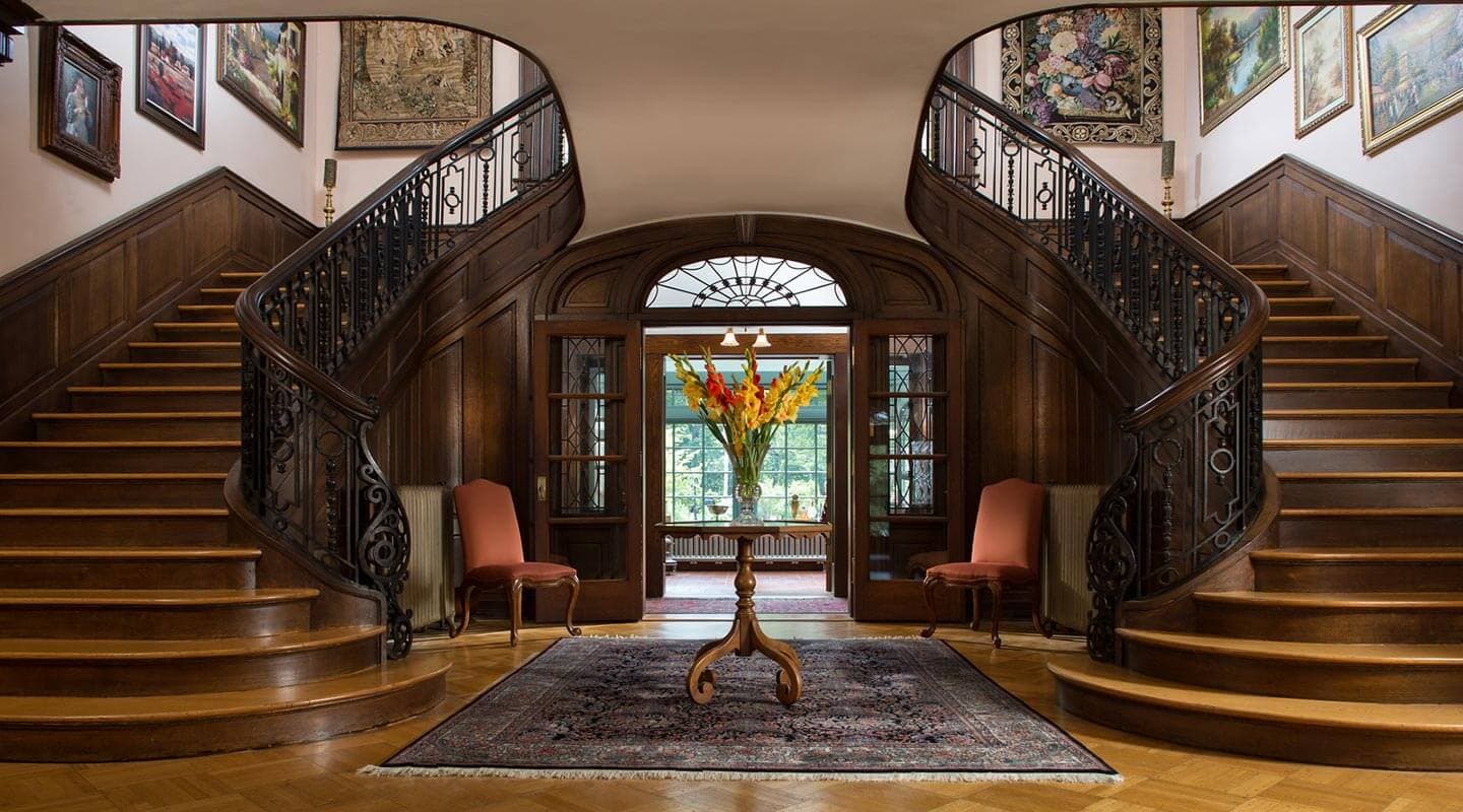 A grand entryway with a double staircase, antique decorations, and a floral centerpiece on an ornate rug.