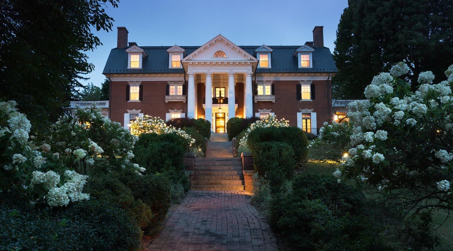 A grand brick mansion with white columns, illuminated gardens, and a pathway leading to the entrance at dusk.