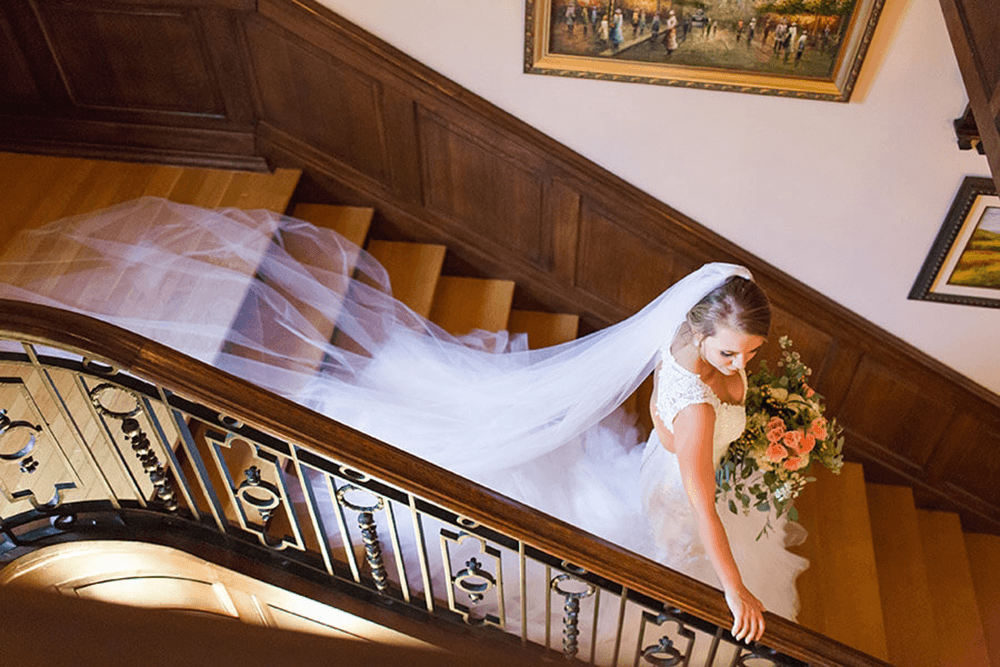A bride descends a staircase, holding a bouquet and wearing an elegant wedding dress with a flowing veil.