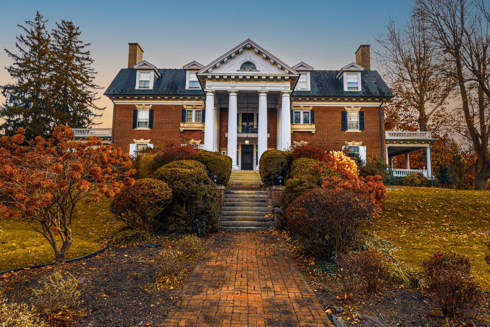 A grand brick mansion with white columns surrounded by autumn foliage and a brick pathway.